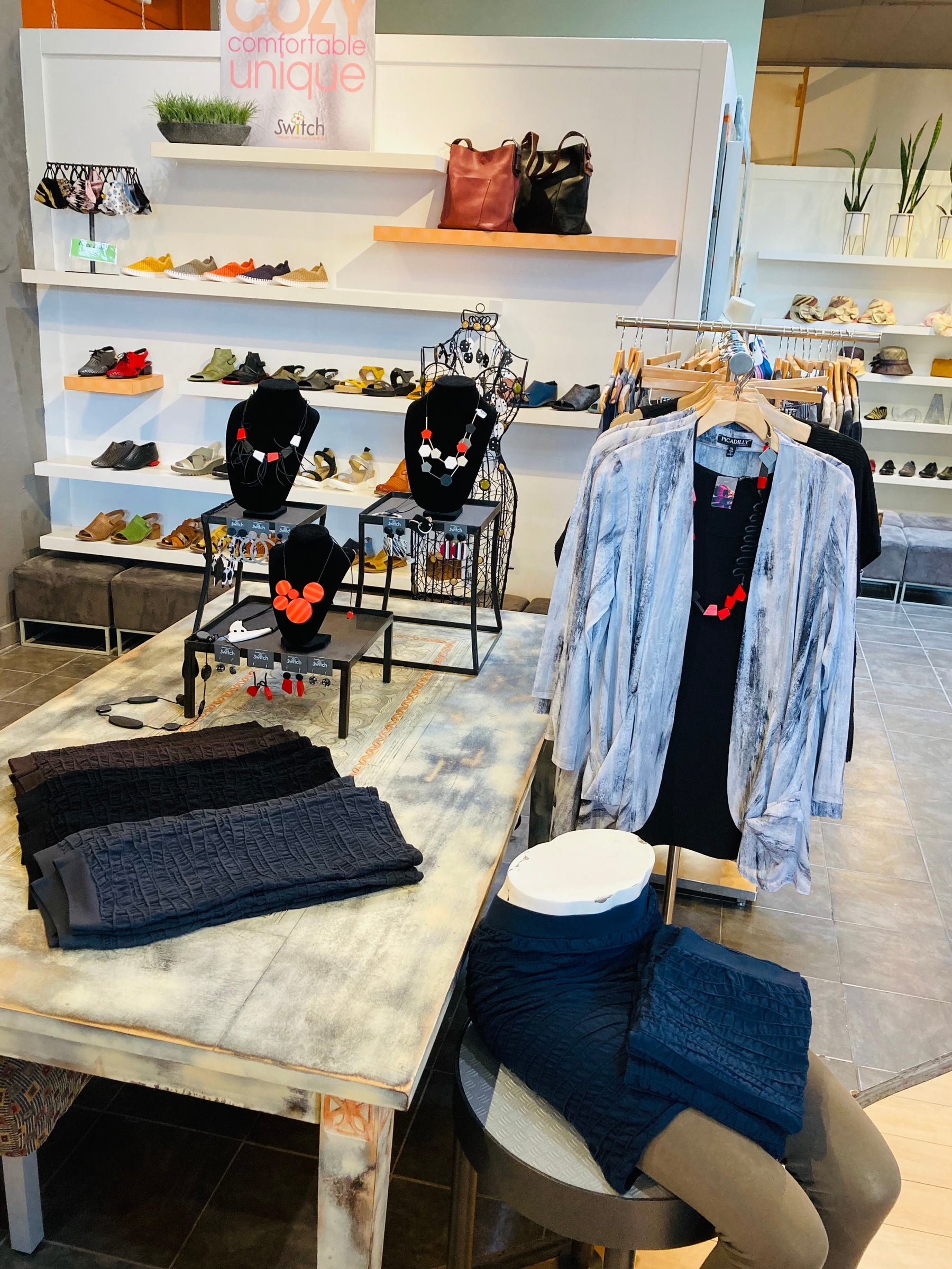 A table with skirts and jewelry sits in the middle of the store in front of a wall of colorful shoes.